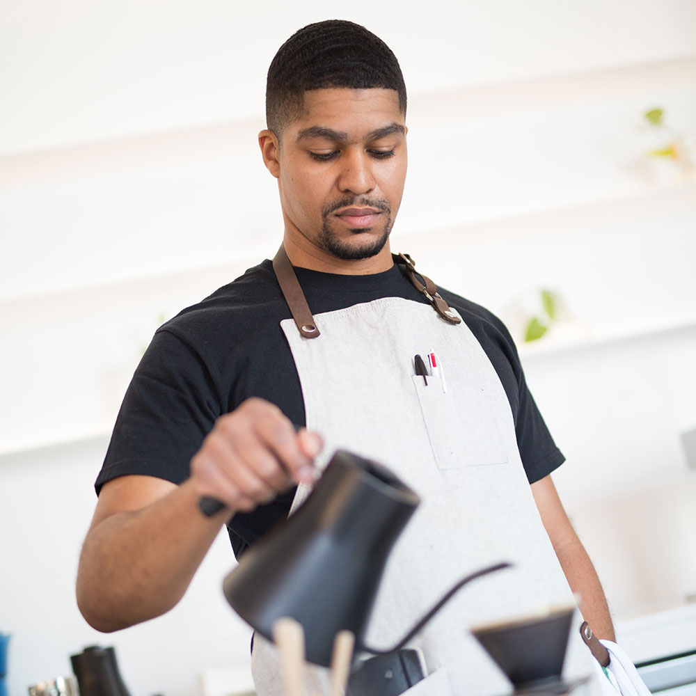 Male employee wearing a white apron in a modern kitchen slow dripping coffee at a cafe in Wake County