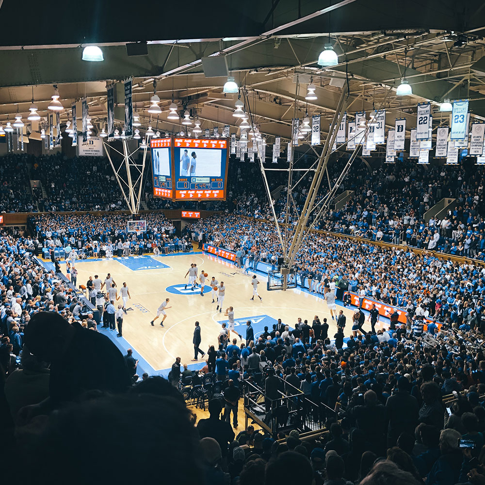 A large crowd at an indoor basketball stadium with retired blue and white jersesys hanging from the ceiling at Duke University in Wake County