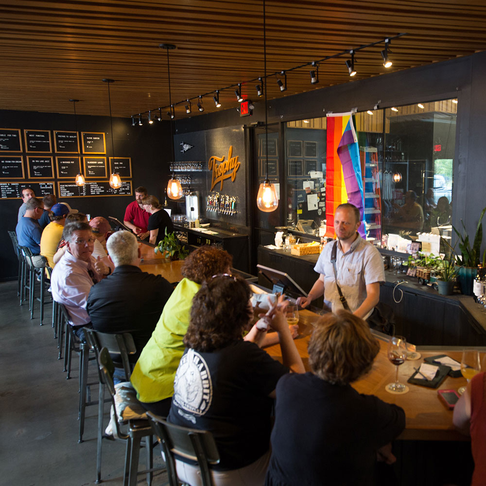 Aerial view of an indoor bar with patrons conversing and a bartender standing in front of a pride flag in Wake County