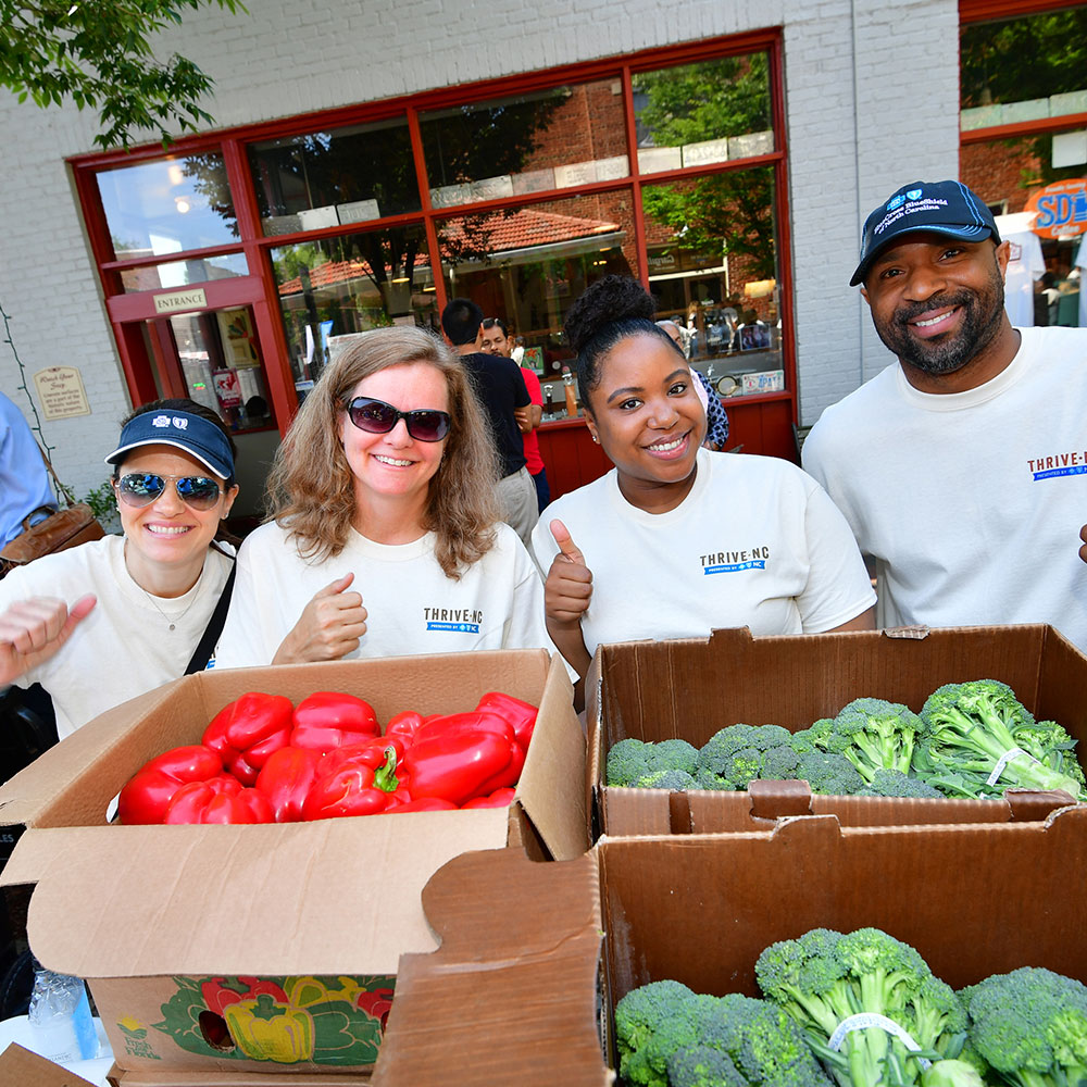Four individuals smiling and putting a thumbs up behind boxes of fresh produce in Wake County