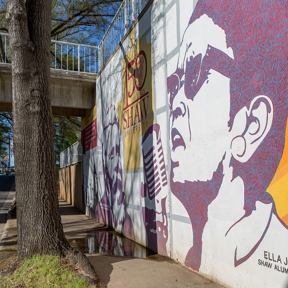 A black, white, orange, red and green mural of Ella Josephine Baker wearing sunglasses on Shaw University's campus near a bridge and main road in Wake County