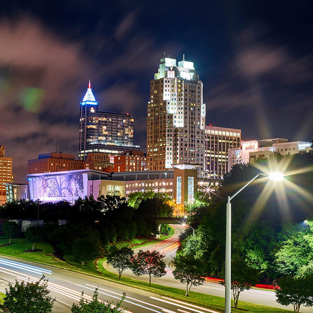 An aerial view of Raleigh at night with the convention center, a few skyscrapers, and a well lit interstate in Wake County