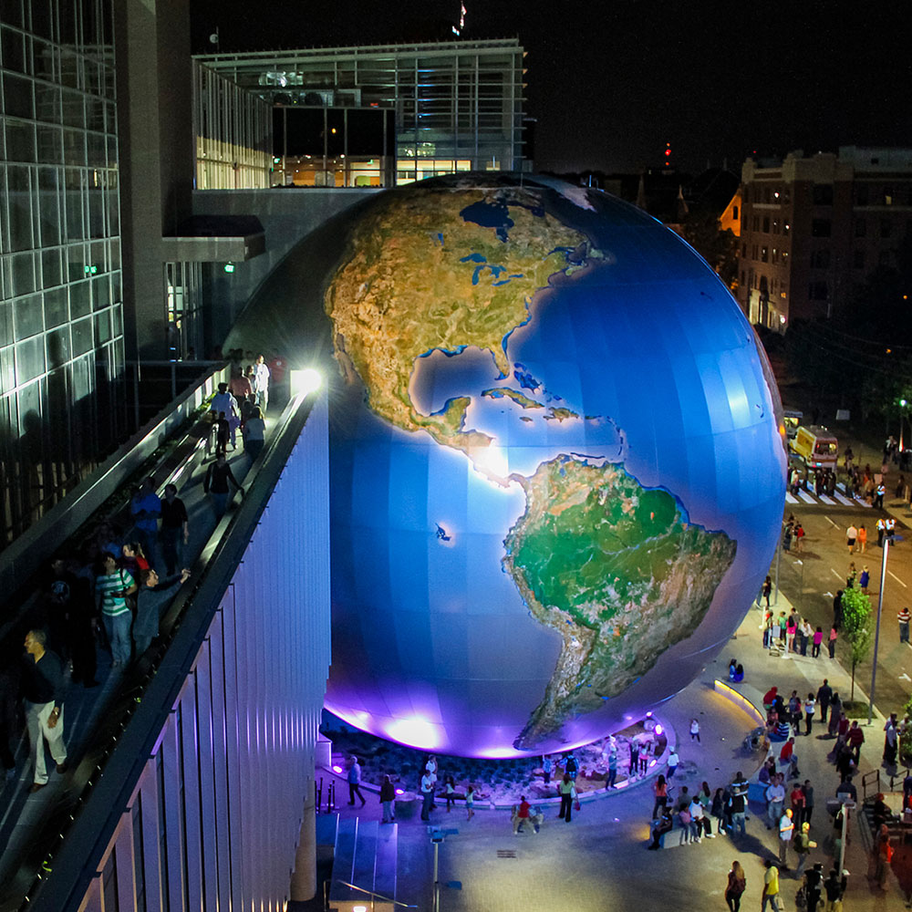A large model of the globe stands illuminated in a bustling downtown city setting with lots of people gathered around taking pictures in Wake County