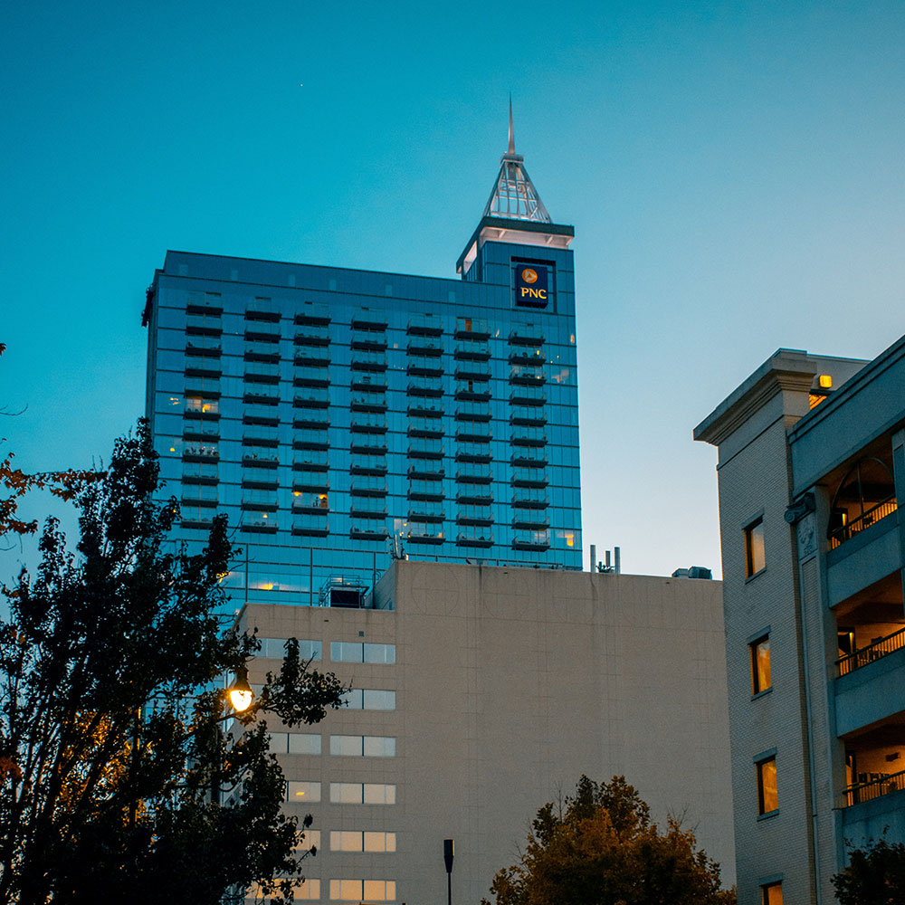 A skyscraper and a large white building in downtown Raleigh with string lights and a law enforcement vehicle in the distance in Wake County