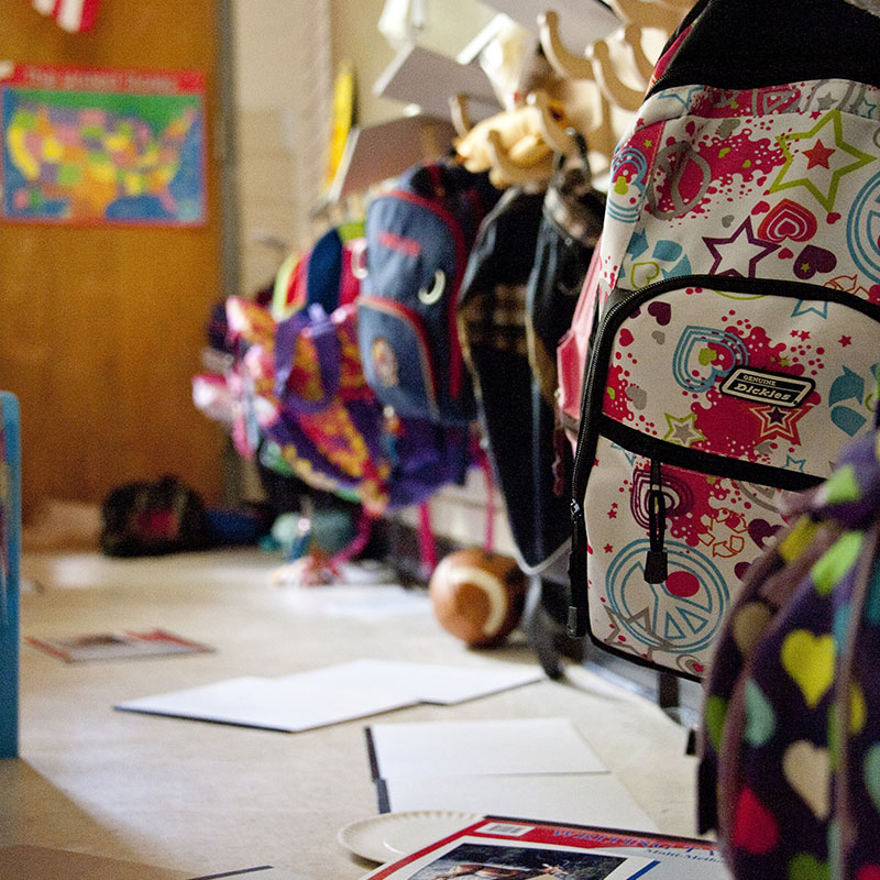 Colorful elementary school backpacks are hung on hooks with scattered papers and coloring pencils beneath them in Wake County