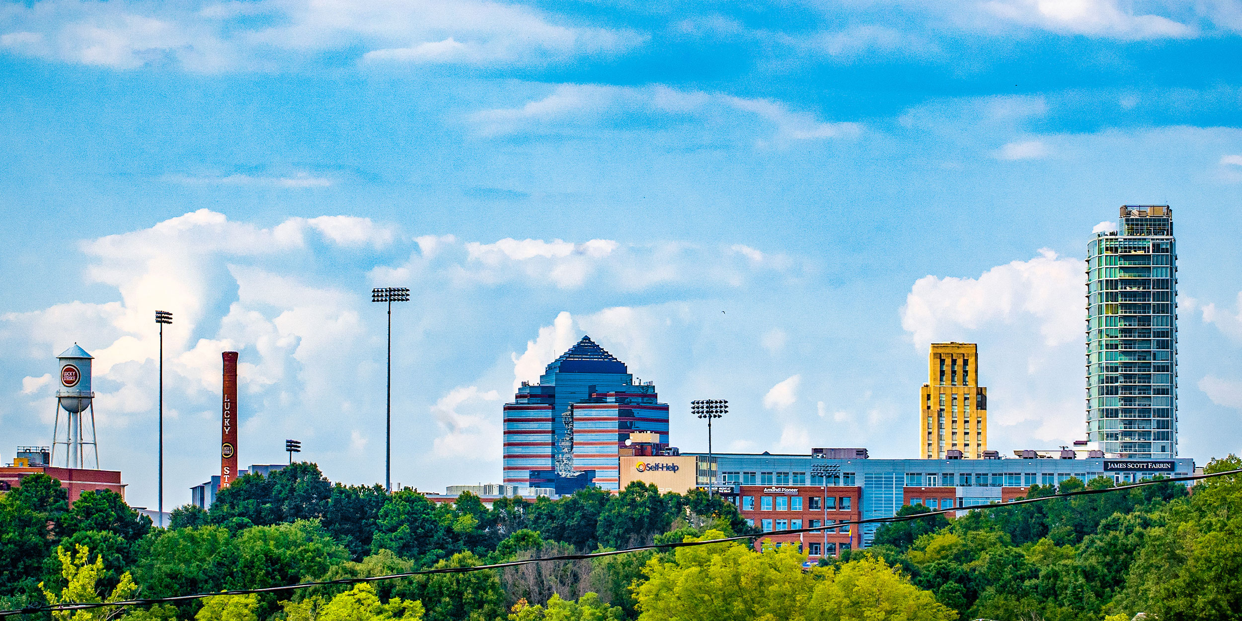 The downtown Durham skyline, with a few tall buildings, a Lucky Strike water tower, and tall industrial lights with lush green spaces in Wake County