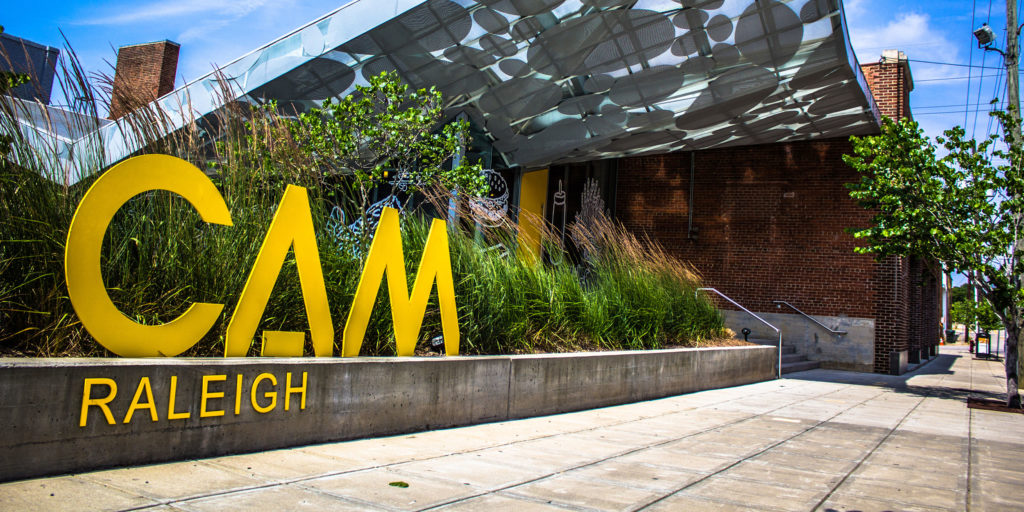 A sign for the CAM Raleigh outside of a curved glass archway and brick building in Wake County