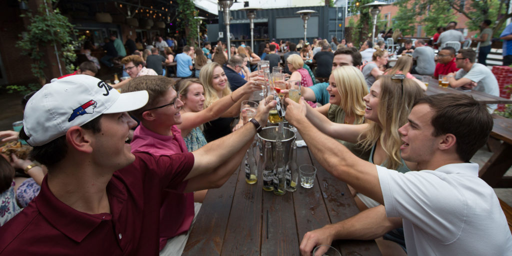 Group of friends at a long rectangular table at an outdoor venue raise their drinks together in the center in Wake County