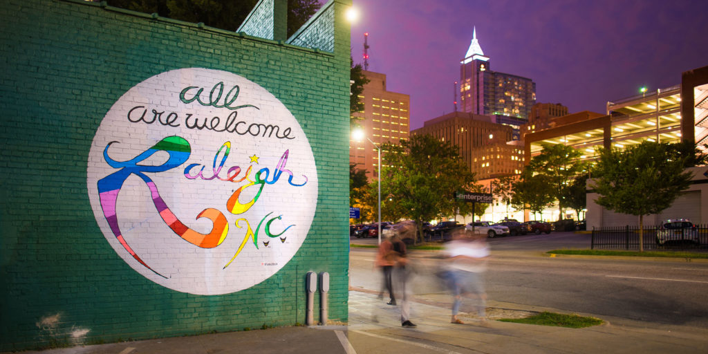 A green painted brick wall with a mural that says All are welcome to Raleigh with rainbow letter with the skyline in the background in Wake County