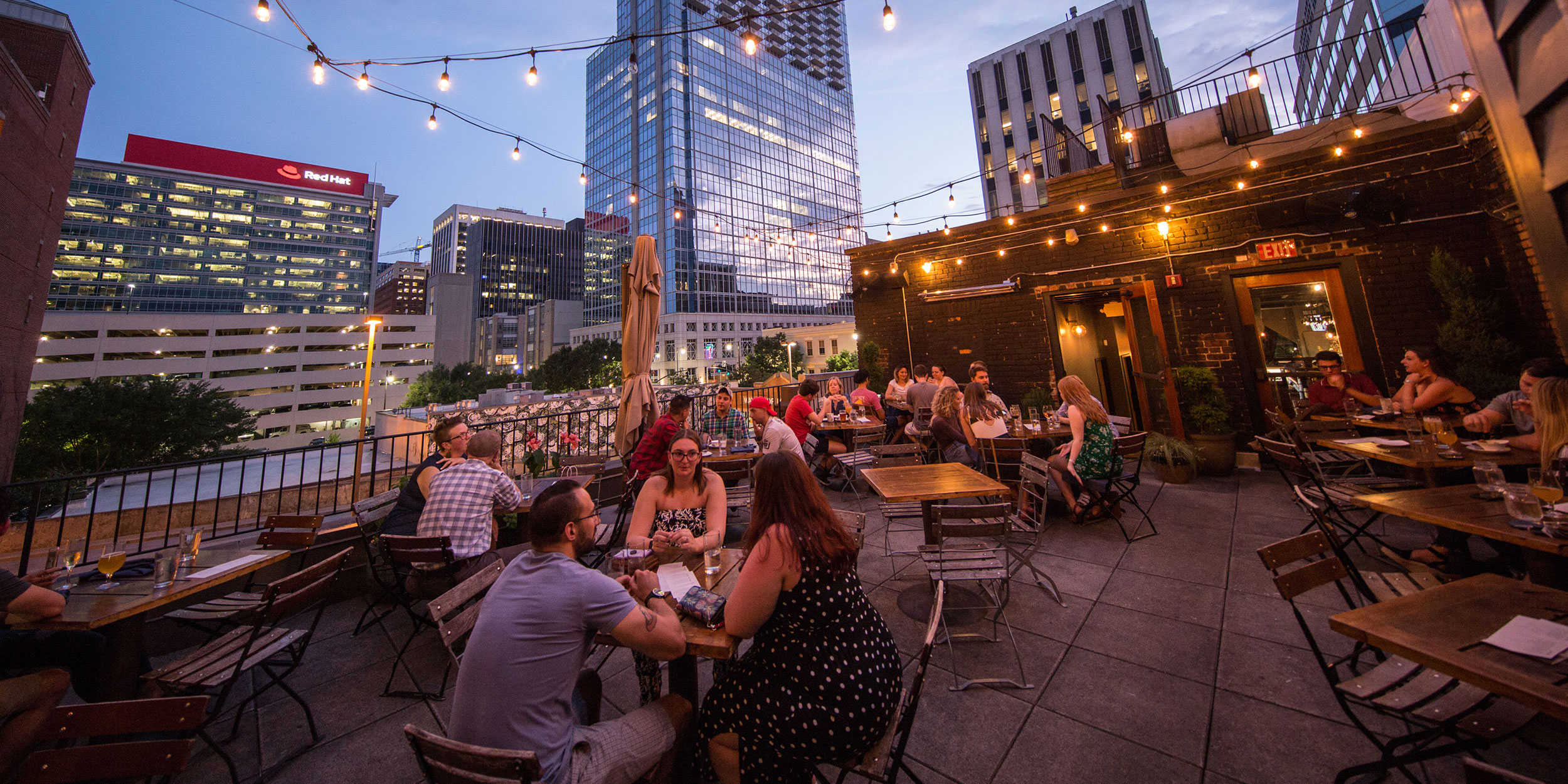 Groups of people sit at an outdoor rooftop restaurant at night with hanging lights and the downtown city skyline behind them in Wake County