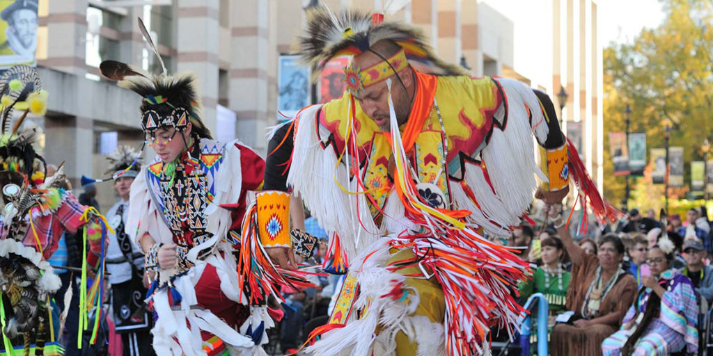 Ceremony of individuals dressed in traditional Native American uniform and headdresses with an onlooking crowd in Wake County