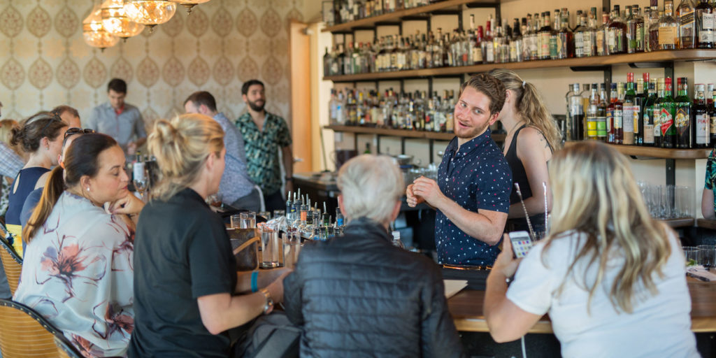 A crowd gathers at a bright, well lit bar with a male bartender talking to a female guest with shelves stocked full of various alcohols in Wake County