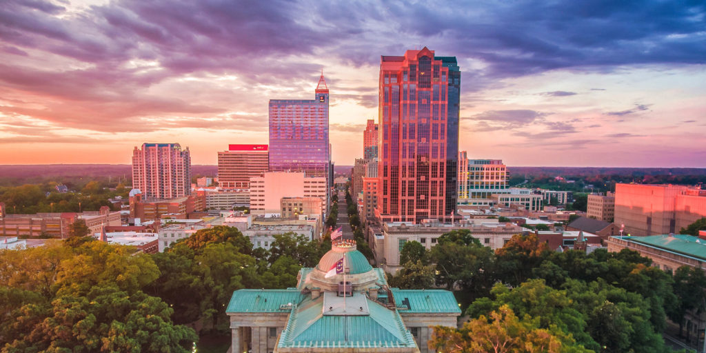 An aerial shot of a downtown city at sunset with a stone and bright green roofed government building and skyscapers in the background in Wake County