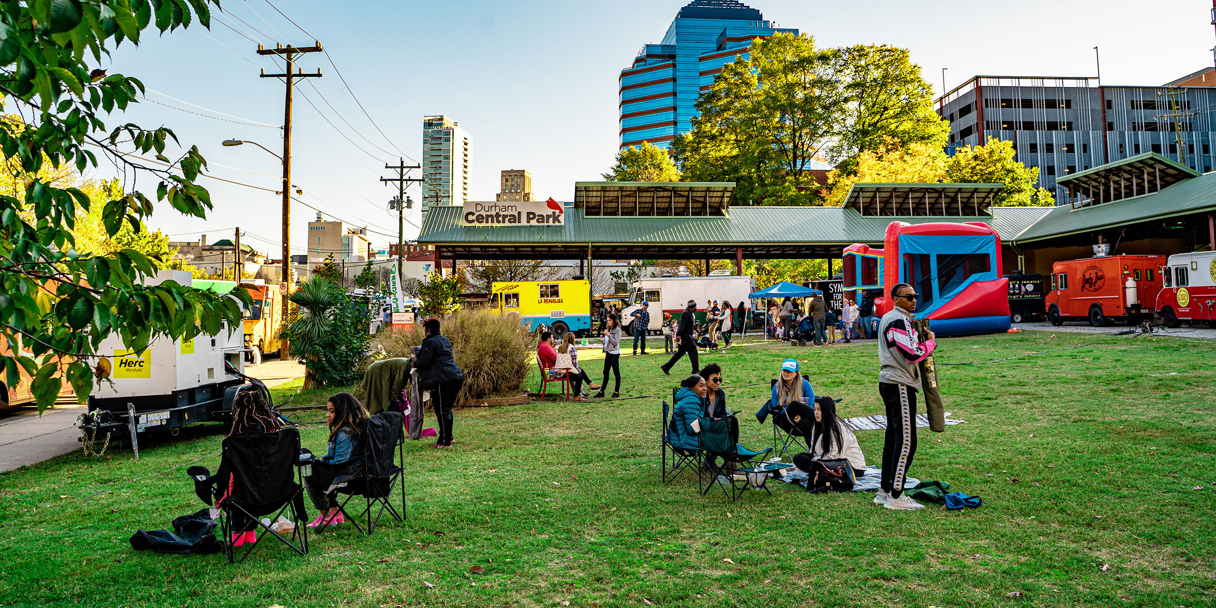 A few clusters of people in lawn chairs sitting in groups on a green space surrounded by food trucks with the Durham skyline in Durham, NC