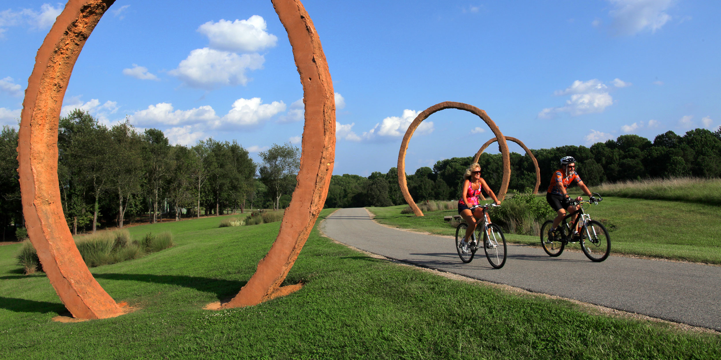 A couple ride bikes alongside each other at a lush park with large sculptures of arches in Wake County