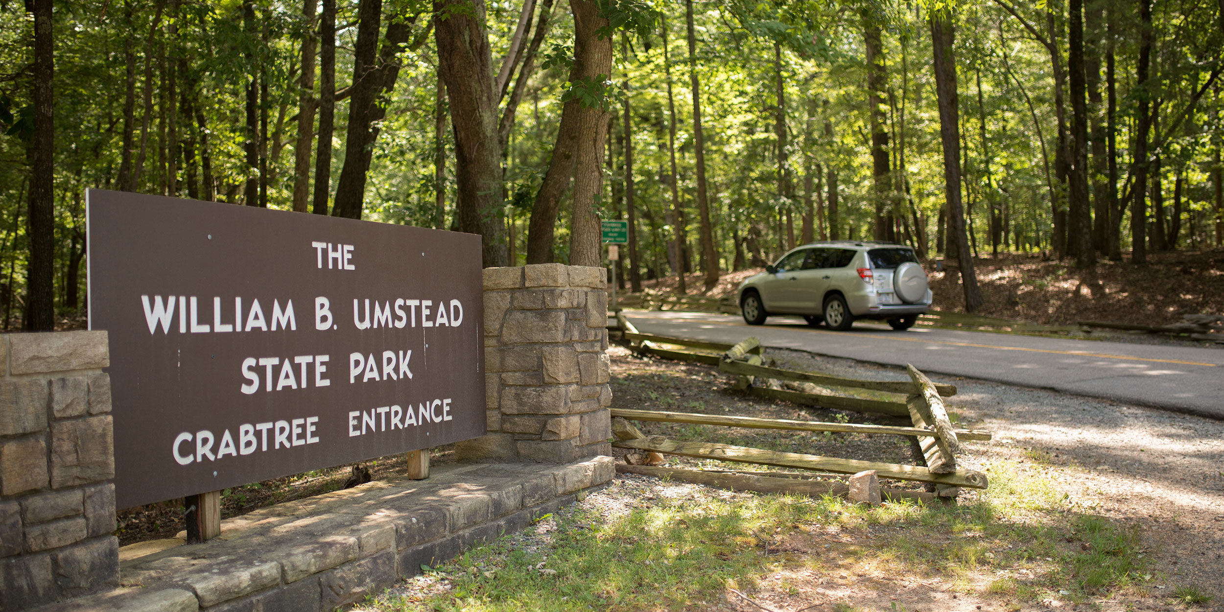 Entrance sign to William B Umstead State Park Crab Tree entrance with trees and a car in the background in Wake County