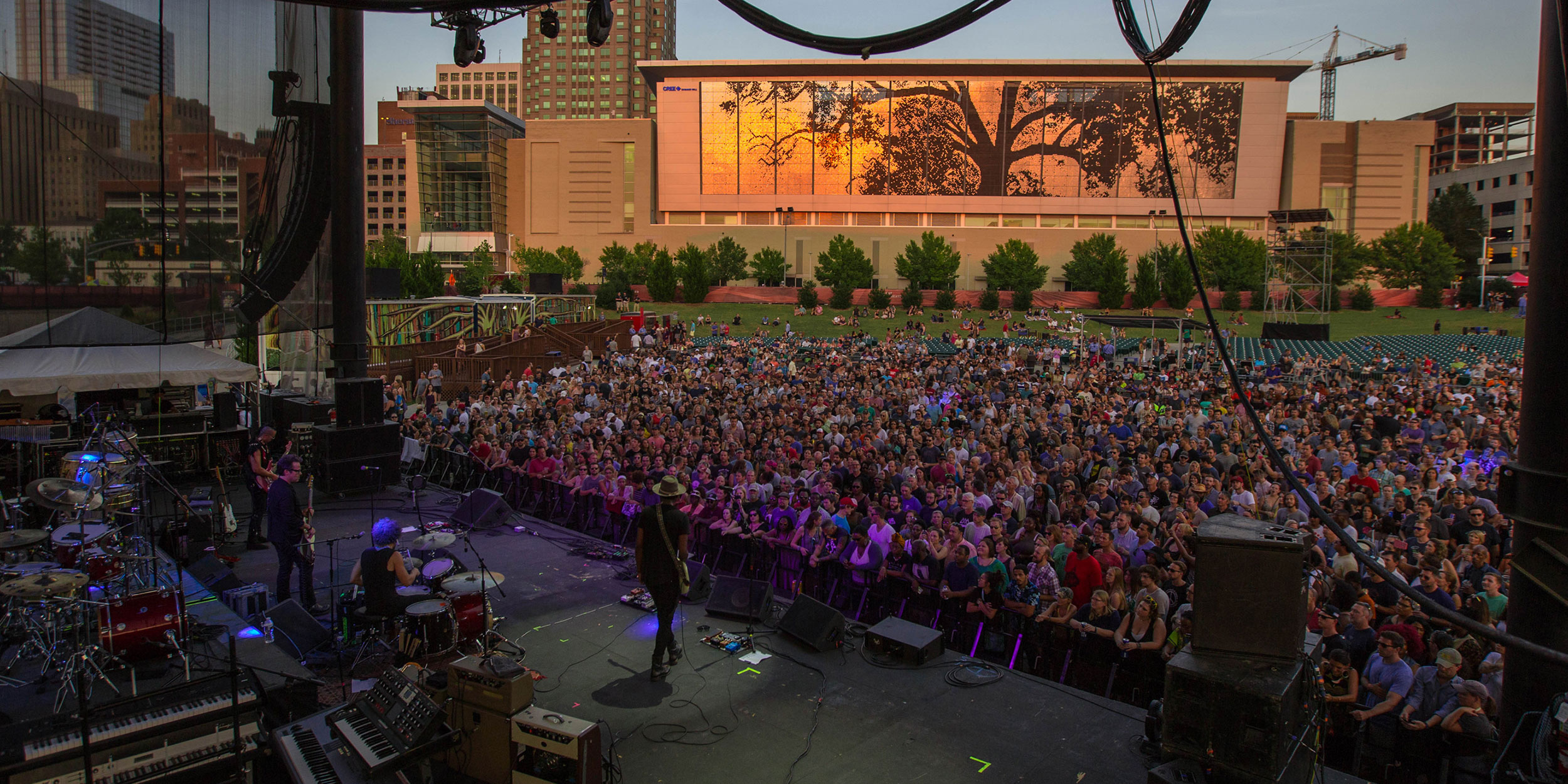 Overlooking a group of performers on stage at a concert with a large crowd and a city skyline in the background at sunset in Wake County