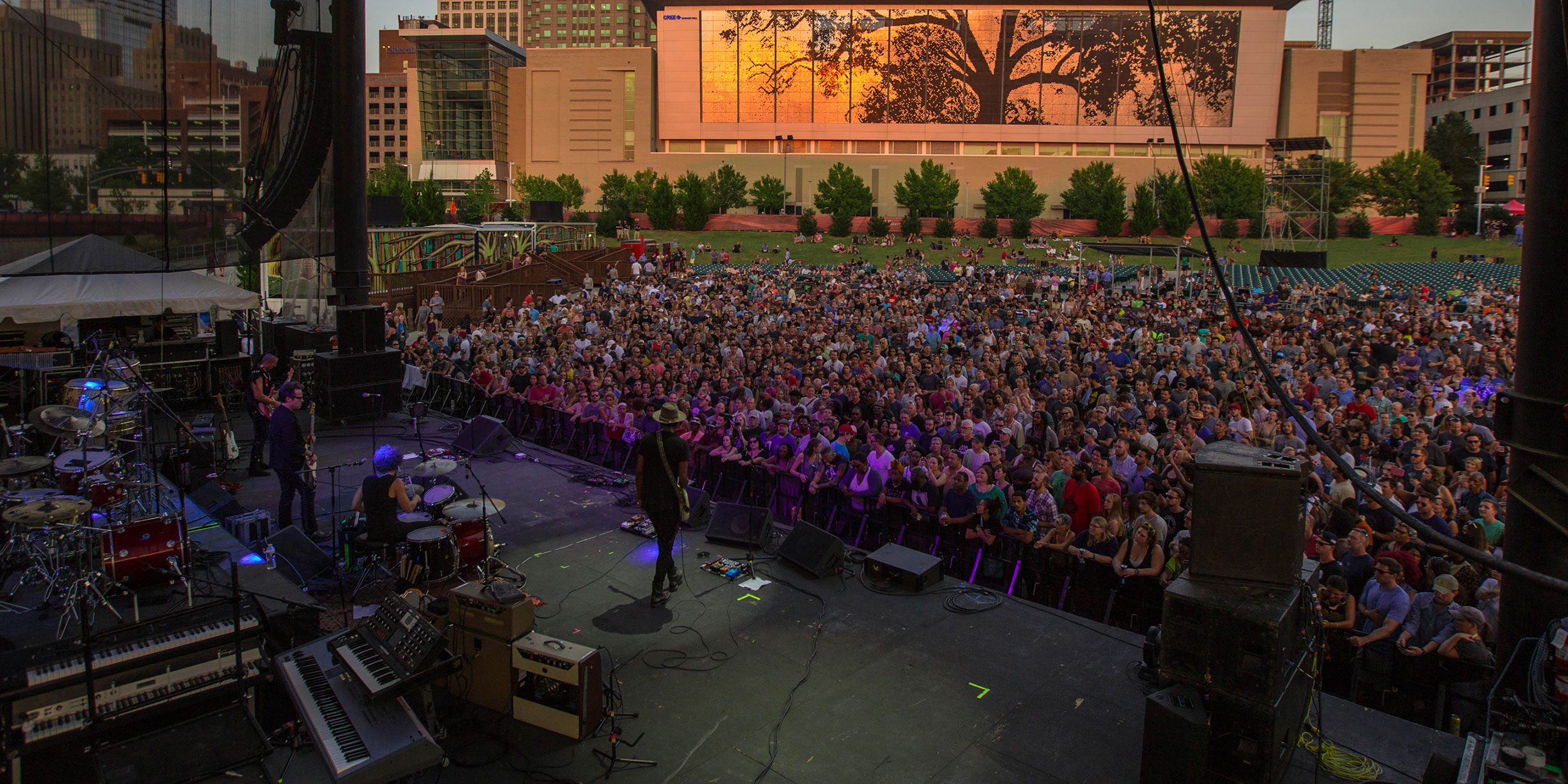 Overlooking a group of performers on stage at a concert with a large crowd and a city skyline in the background at sunset in Wake County