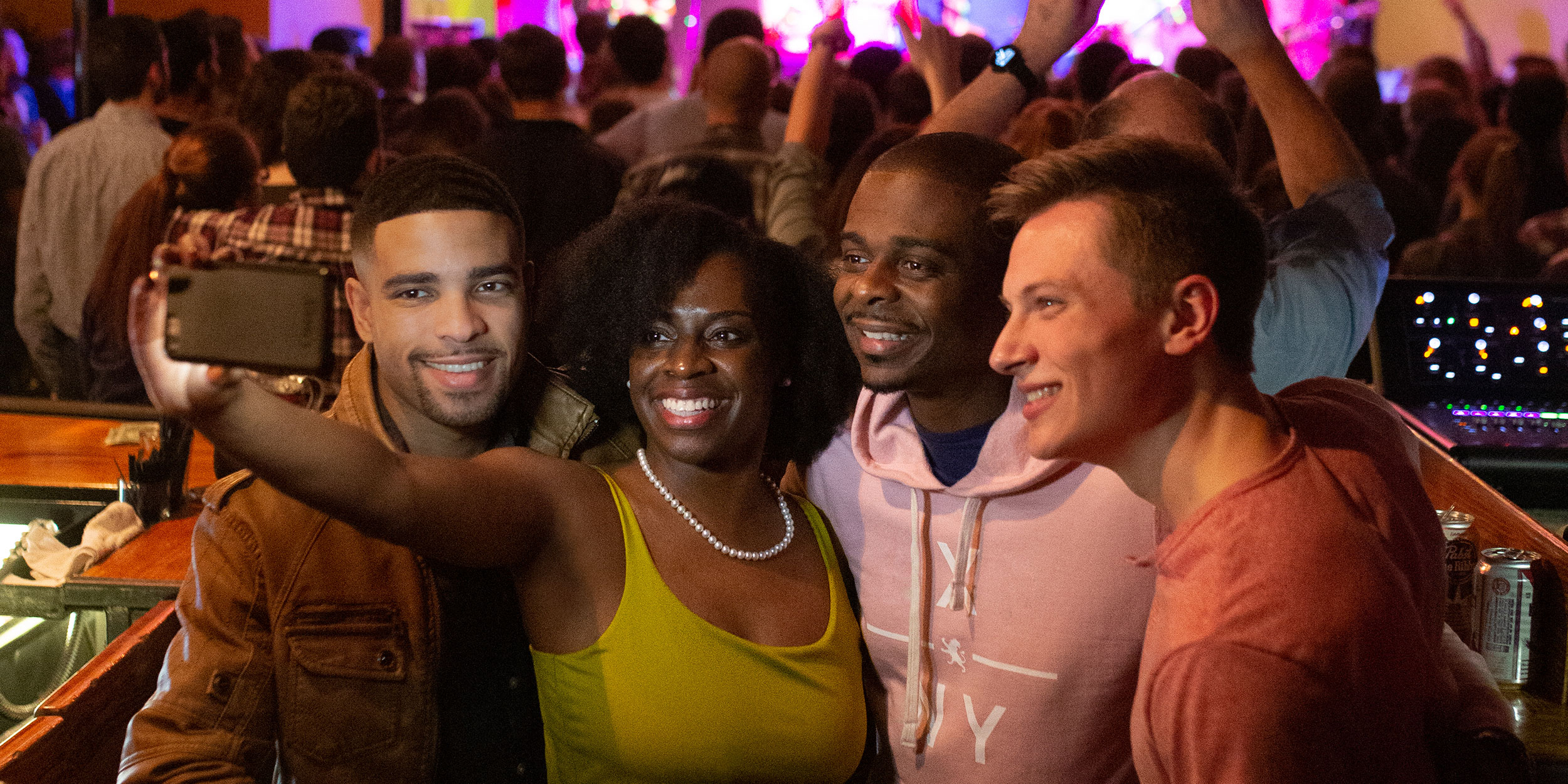 A group of friends take a selfie in an indoor concert hall with a large crowd behind them and strung colored lights in Wake County