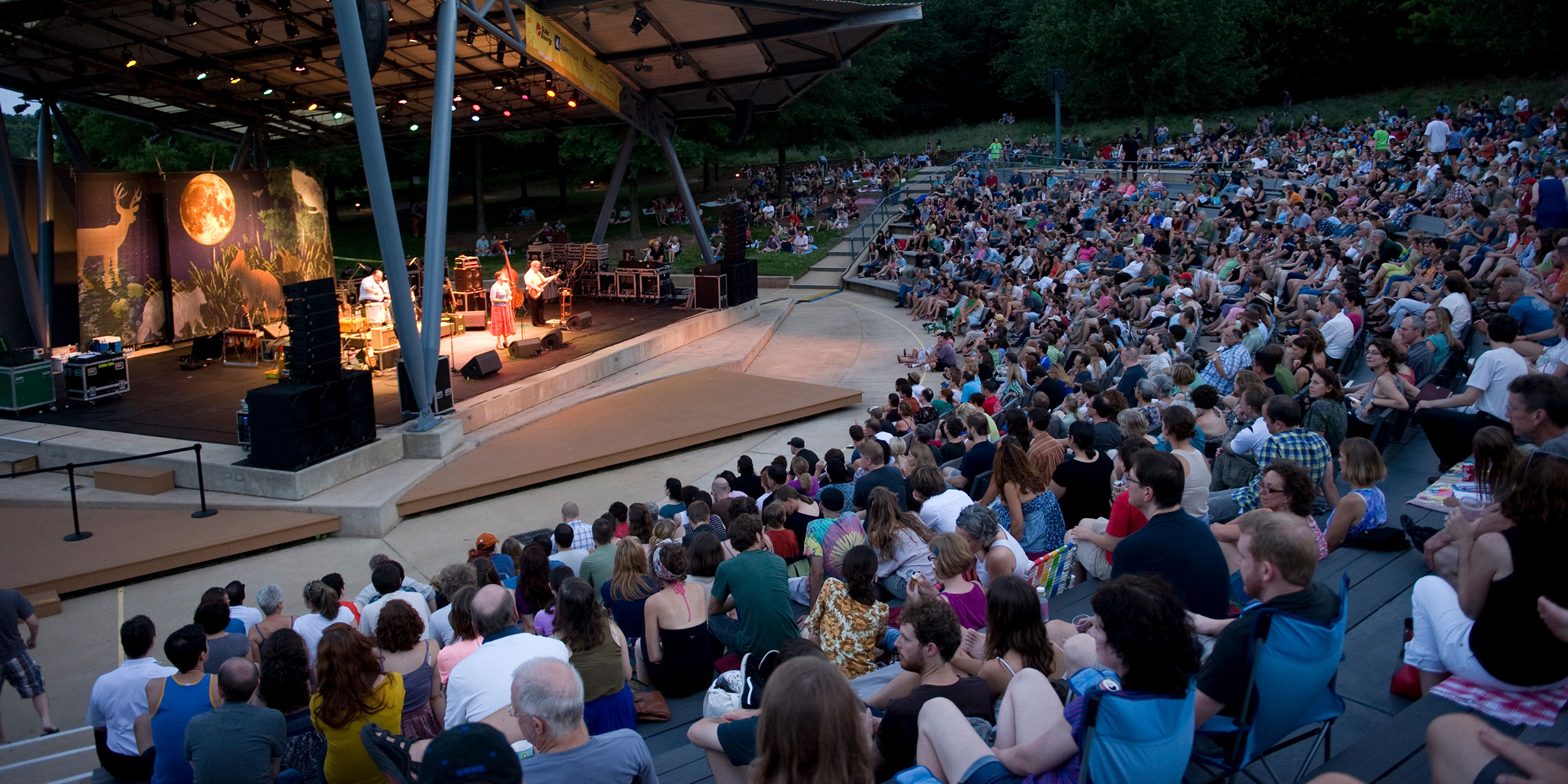 Large crowd gathered outside an outdoor ampitheatre with a group on stage in front of a painted backdrop of forest animals in Wake County