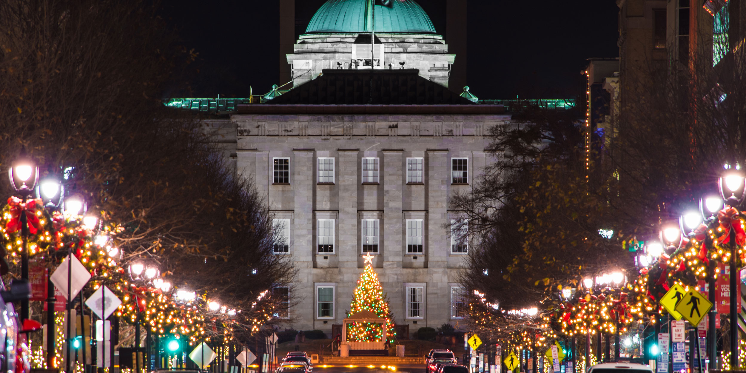 Lit up holiday tree in the distance of a lit street with winter holiday decorations on light poles and a well lit dome building in Wake County