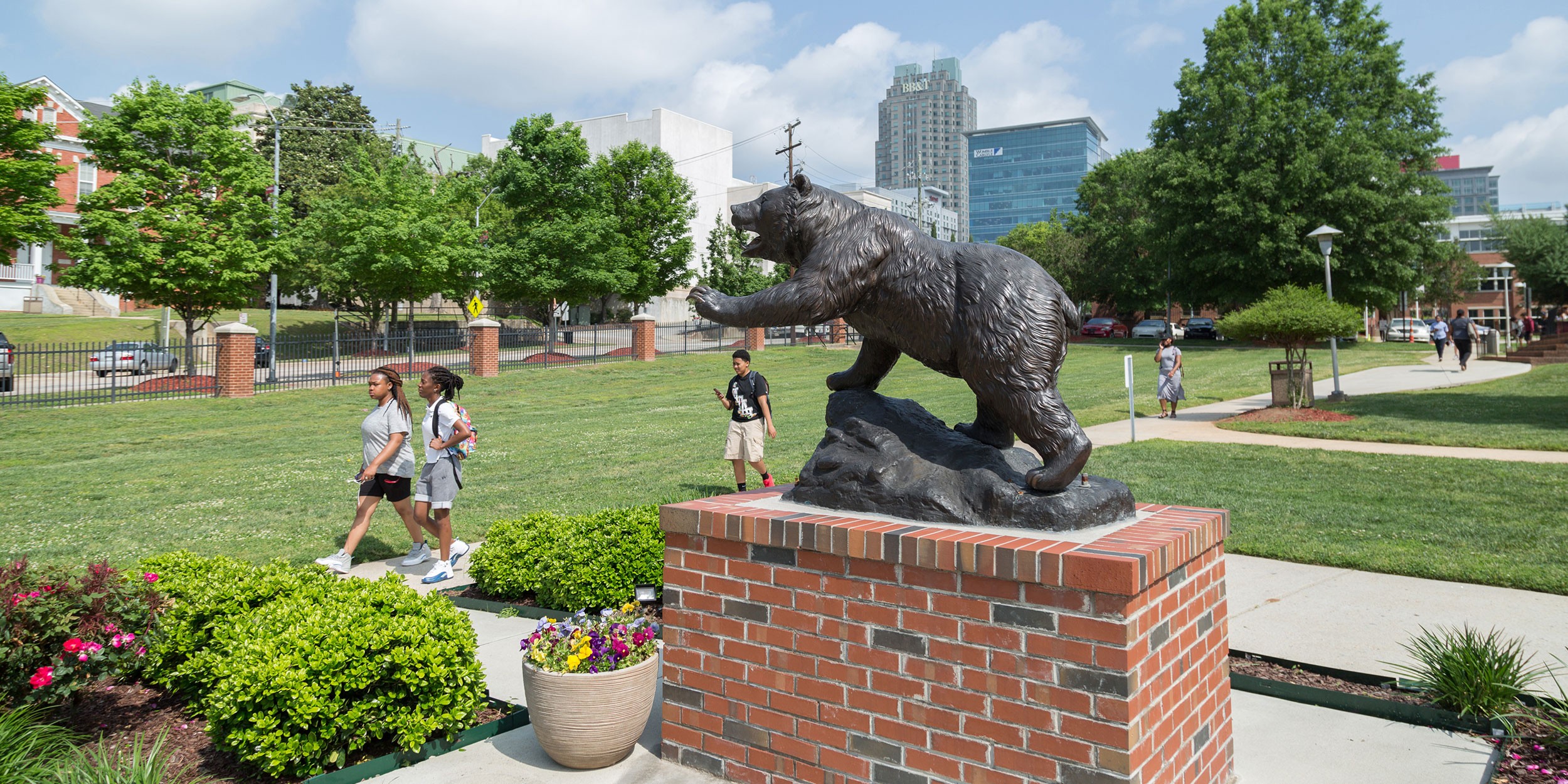 Two girls in the distance walk on Shaw University campus passing a statue of a bear with the city skyline in the backdrop in Wake County