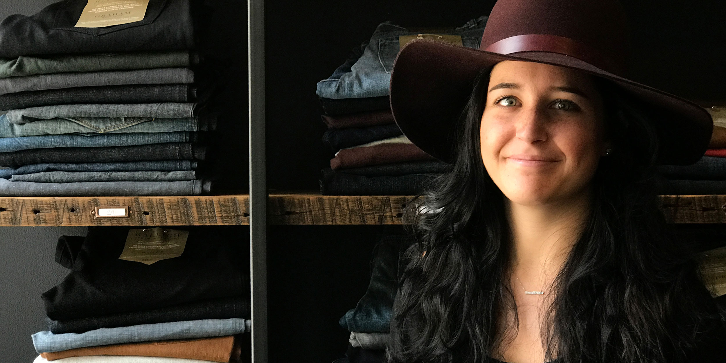 A young woman wearing a large brimmed hat in front of shelves filled with folded jeans in Wake County