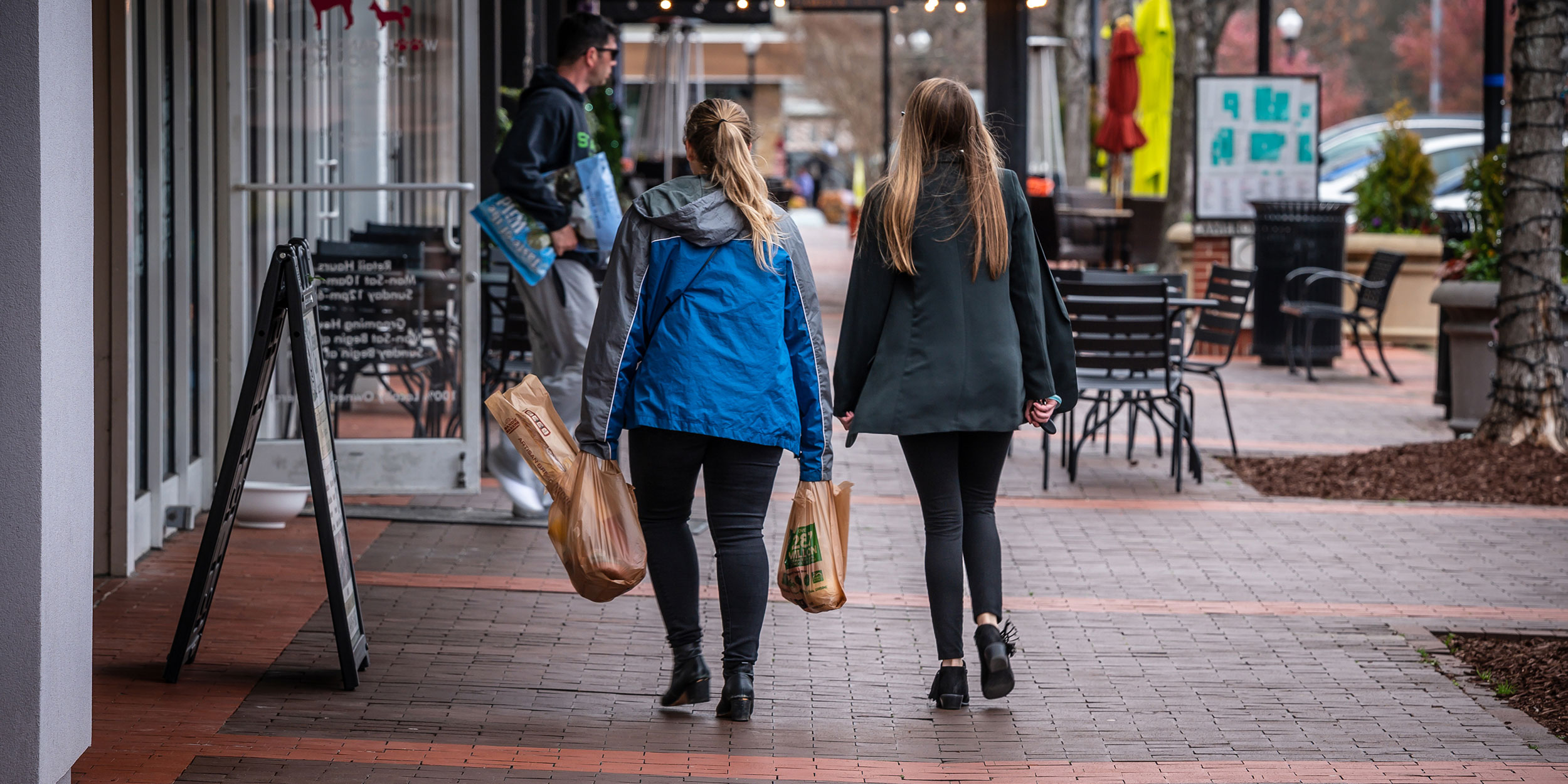 Two girls walk in the Village District holding bags of groceries while passing store fronts in Wake County