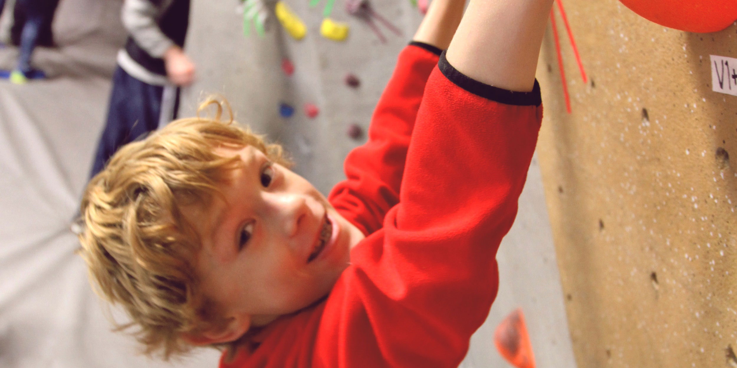 Child smiling and hanging on to a rock at at the Triangle Rock Club, an indoor rock climbing facility in Wake County