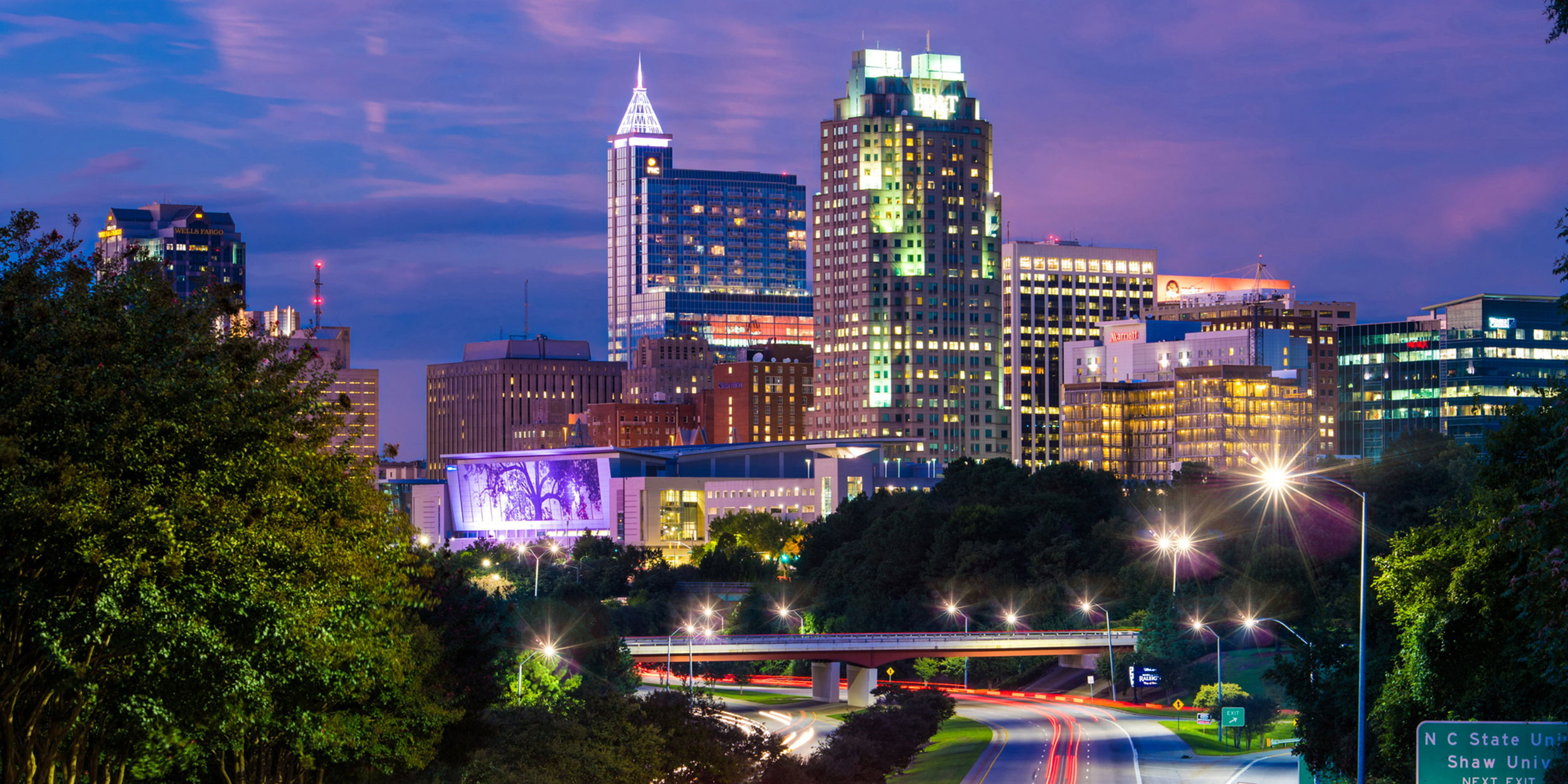 City skyline with a purple sky at night with lit buildings and over exposed car lights running through a major highway in Wake County