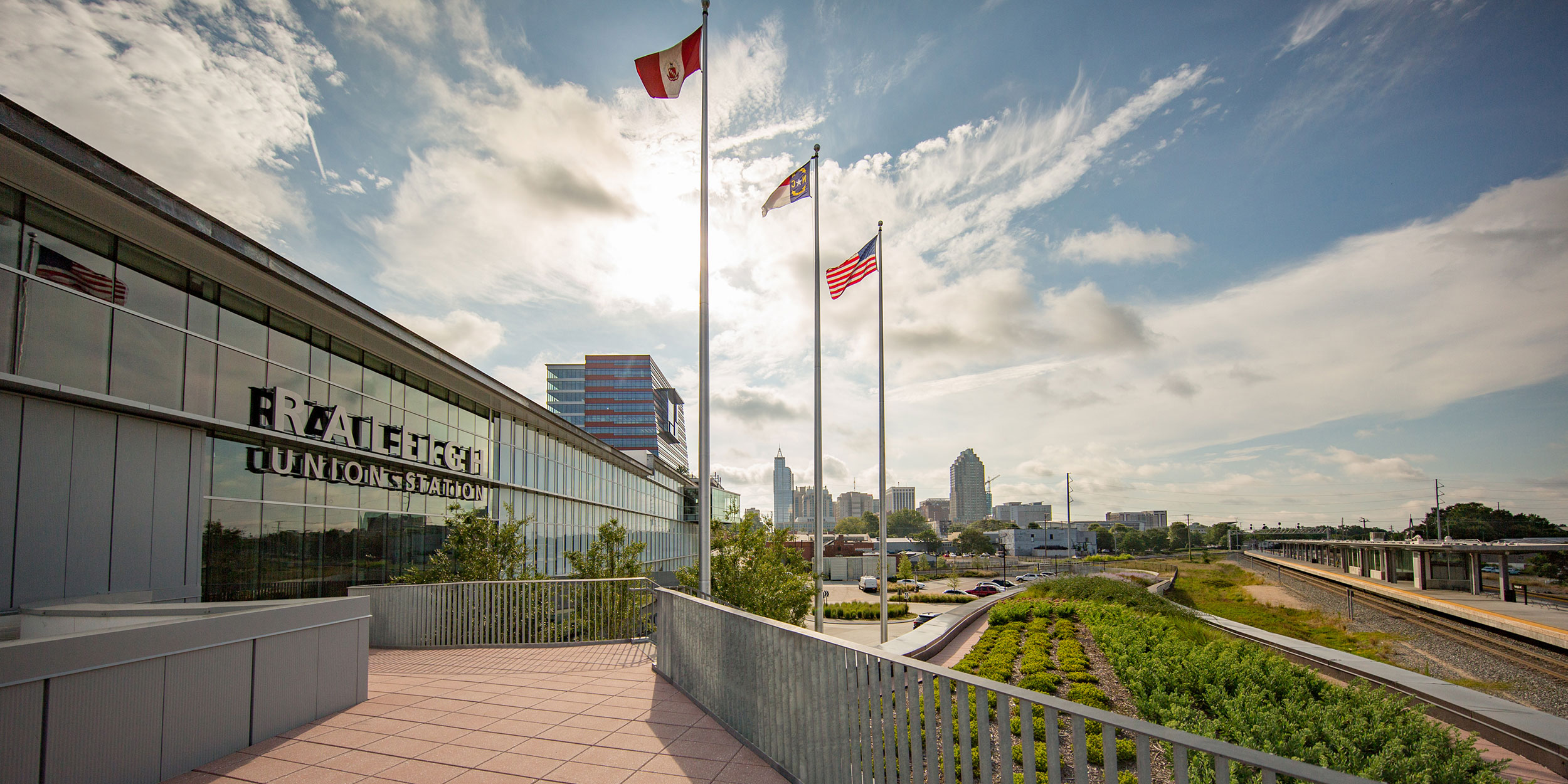 Scenic photo of a glass building with three flag poles in front, metro tracks in the distance and the Raleigh skyline in Wake County