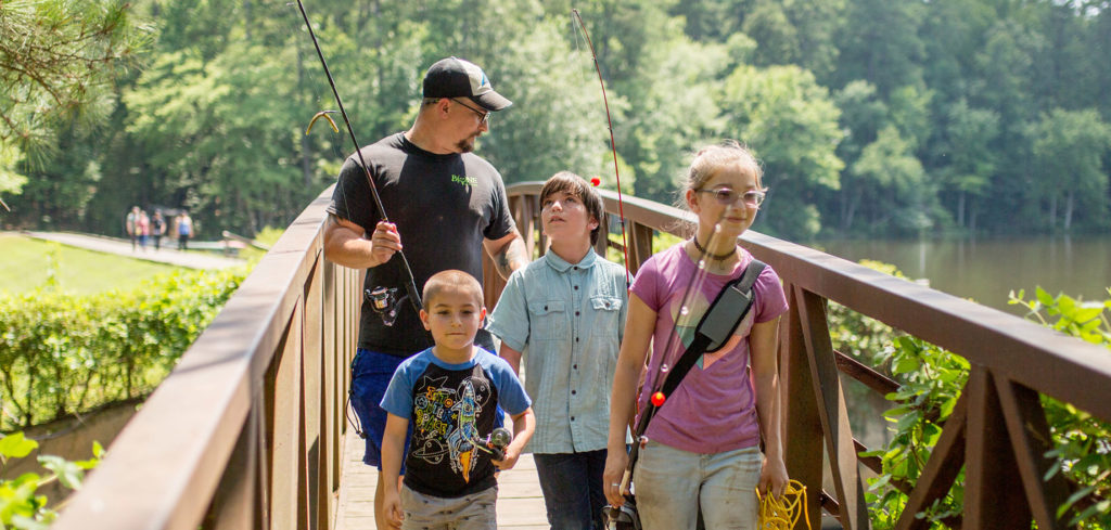 A man and three children walk across a wooden bridge holding fishing gear with a river and scenic foresty in the background in Wake County
